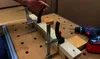 A person's hand is shown clamping two pieces of wood together on a workbench. A drill is visible in the background. Náhľad obrázku produktu
