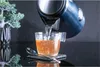 Water being poured from a kettle into a glass cup with a spoon next to it, with a decorative plant in the background. Podgląd zdjęcia produktu