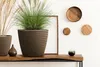 A brown knitted-style planter with a green plant sits on a wooden shelf, next to dried grasses and decorative wooden circles on a white wall. Podgląd zdjęcia produktu