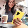 A woman is holding a yellow phone case and a coffee cup at a table with a laptop. Produktbild-Vorschau
