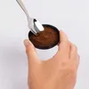 A hand holding a small black container filled with coffee grounds, with a spoon scooping some of the grounds. Produktbild-Vorschau