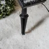A close-up view of a black stake for a solar light, partially inserted into artificial snow, with a hint of a green bush in the background. Produktbild-Vorschau