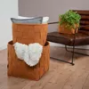 Two woven felt storage baskets are stacked on a wooden floor. The bottom basket contains a white fluffy blanket. The top basket contains folded gray and white towels. A brown leather chair with a potted plant is visible in the background. Anteprima dell’immagine prodotto