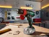 A cordless drill is shown on a workbench in a workshop setting, with tools and wood pieces around it. Podgląd zdjęcia produktu