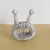 A gray cosmetic headband with two plush eyes on stalks is displayed on a clear stand against a white and tan background. Produktbild-Vorschau