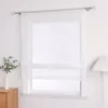 A white sheer Roman blind is shown hanging on a window, with a small plant in a vase on a wooden cabinet to the left, and a floor lamp with a pleated shade to the right. Produktbild-Vorschau