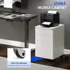 A printer sits on top of a white filing cabinet with three drawers, next to a standing desk. A close-up inset shows the anti-tip wheels of the filing cabinet. Anteprima dell’immagine prodotto