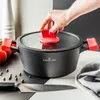 A hand is shown placing a red lid onto a black pot with red handles. The pot is on a countertop with tomatoes, peppercorns, and a knife. Podgląd zdjęcia produktu