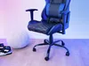 A blue and black gaming chair is shown in a room with a white pouf and sneakers. Produktbild-Vorschau