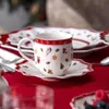 A festive Christmas-themed mug and saucer set on a red tablecloth, with other plates in the background. Náhled obrázku produktu