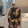 A young boy is holding up a blanket with a firefighter's uniform printed on it, standing in a bedroom next to a bed and a dresser with a TV. Produktbild-Vorschau