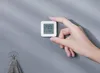 A hand holding a small white digital thermometer against a gray wall. Podgląd zdjęcia produktu