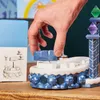 A hand is placing a blue building block onto a toy structure. The structure appears to be part of a game or playset with colorful gem-like pieces on top of a tower. Podgląd zdjęcia produktu