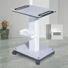 A white cosmetic trolley with a tray and wheels, shown in a studio setting with a potted plant in the background. Náhled obrázku produktu