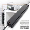 A black power strip is shown in a close-up, angled view, with a smaller black power strip with plugs visible on the left. The background is a white brick wall and a white shelf with a toaster and a plant. Podgląd zdjęcia produktu