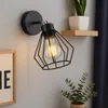 A modern black wall lamp with a geometric cage design is mounted on a gray wall, illuminating a light bulb. A framed picture and a potted plant are visible nearby. Produktbild-Vorschau