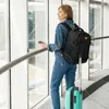 A woman stands in a modern, well-lit hallway, possibly an airport or train station, looking out a large window. She is wearing a denim jacket and jeans, carrying a black backpack, and has a turquoise suitcase next to her. She holds a passport in her hand. Produktbild-Vorschau