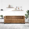 A brown Chesterfield sofa is shown in a well-lit room with shelves on the wall and a plant in the corner. Náhled obrázku produktu