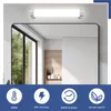A bathroom mirror with a light fixture above it, reflecting a window and some shelves with plants and bottles. Below the mirror are four circular icons with text. Anteprima dell’immagine prodotto