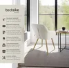 A cream-colored chair with gold legs is placed next to a small round table with a book and a mug, in a well-lit room with large windows overlooking a forest. Produktbild-Vorschau