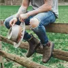 A person sitting on a wooden fence in a grassy field, holding a hat and wearing boots. Náhľad obrázku produktu