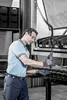 A man in a factory setting wearing work gloves and safety glasses, holding a tool. Produktbild-Vorschau