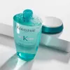 A bottle of Kerastase shampoo with some foam on top, placed on a white surface with a teal and white lid in the background. Podgląd zdjęcia produktu