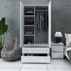 A white wardrobe with two doors and two drawers, partially open, showing clothes and shoes inside. A beanbag chair and a plant are visible to the left, and a nightstand with a lamp is to the right. Produktbild-Vorschau