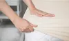 Hands are shown stretching a beige fitted sheet over a mattress. Produktbild-Vorschau