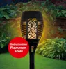 A solar-powered garden torch with an LED flame effect, set in a garden with plants and a stone wall. Produktbild-Vorschau