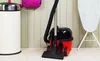 A red and black vacuum cleaner is placed on a light-colored floor next to a white trash can and an ironing board. Náhled obrázku produktu