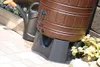 A brown rain barrel on a gray stand next to a black watering can and some plants. Produktbild-Vorschau
