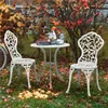 A white metal bistro set for two is set up in a garden with autumn leaves on the ground and trees in the background. Produktbild-Vorschau