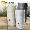 Three KADAX rain barrels in different colors are displayed outdoors on a tiled patio, with greenery in the background. Náhled obrázku produktu