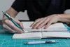 A person is writing in a notebook with a ruler and pen, with another pen in the foreground. Produktbild-Vorschau