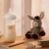 A baby bottle filled with milk sits on a wooden surface next to a stuffed donkey toy and wooden blocks. Anteprima dell’immagine prodotto