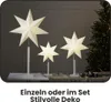 Three illuminated paper star lanterns of varying sizes are displayed on a table in front of a Christmas tree and gifts. Podgląd zdjęcia produktu