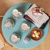 A set of coffee cups and saucers arranged on a blue table, with books and cookies in the background. Produktbild-Vorschau