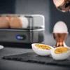 A close-up shot of a boiled egg cut in half, placed on a dark slate, with an egg cooker and an egg cup in the background. Voir les images du produit