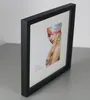 A black picture frame with a white mat and a colorful image of a woman's face is displayed on a gray surface. Produktbild-Vorschau