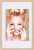 A woman with a large bow hairstyle and vibrant makeup poses with her hands near her face, against a background of large orange polka dots. Produktbild-Vorschau