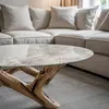 A close-up of a marble and wood coffee table in a living room with a sectional sofa in the background. Podgląd zdjęcia produktu