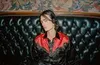 A person wearing headphones sits on a black leather couch in front of a patterned wall. Voir les images du produit