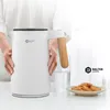 A person is holding a white electric kettle with a wooden handle, with a glass of water and cookies in the background. Produktbild-Vorschau