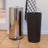 A stainless steel trash can and a black bin are placed on a tiled floor next to a white wall. A checkered towel hangs on the wall behind the bins. Produktbild-Vorschau