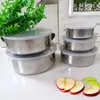 A set of stainless steel food containers with clear lids, stacked on a white surface, with a sprig of green leaves and apple slices in the foreground. Produktbild-Vorschau