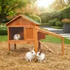 Two rabbits are sitting on straw in front of a wooden hutch with a green roof. A guinea pig is visible inside the hutch. Produktbild-Vorschau