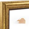 A close-up view of a gold antique-style picture frame with a small wooden sample piece in the foreground. Produktbild-Vorschau