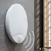 A white motion-sensing outdoor light fixture mounted on a gray tiled wall, with sunlight casting shadows through a window in the background. Produktbild-Vorschau