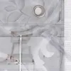 A close-up view of a gray sheer curtain with a leaf pattern and a metal grommet. The curtain is layered over a blurred image of furniture and a rolled rug. Produktbild-Vorschau